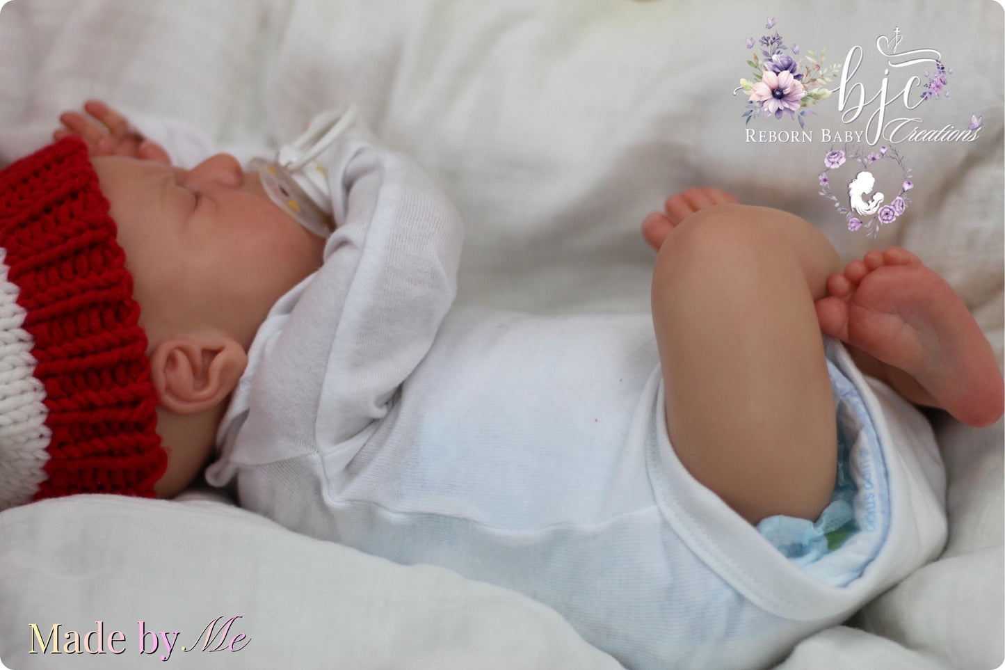 A newborn baby wearing a white onesie and a red knit hat is peacefully sleeping on a white blanket.