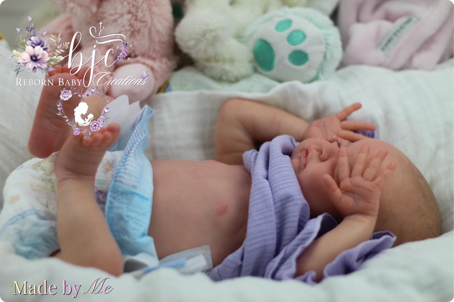 A newborn baby lying on a bed, wearing a purple blanket and surrounded by stuffed animals.